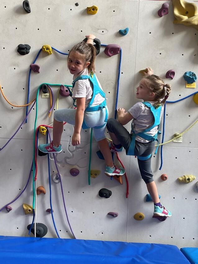 Drei Kinder klettern an einer Kletterwand mit bunten Haltegriffen und Sicherheitsgurten, Matten darunter.