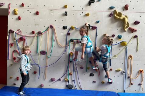 Drei Kinder klettern an einer Kletterwand mit bunten Haltegriffen und Sicherheitsgurten, Matten darunter.