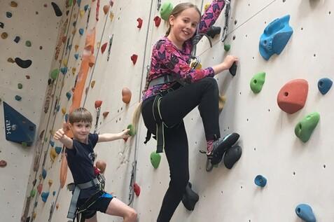Zwei Kinder klettern an einer Kletterwand mit bunten Griffen, lächelnd und in Sicherungsgurten.