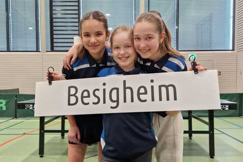 Drei Mädchen in Sportkleidung halten ein Schild mit der Aufschrift „Besigheim“ vor einem Tischtennistisch und Punkteständen.