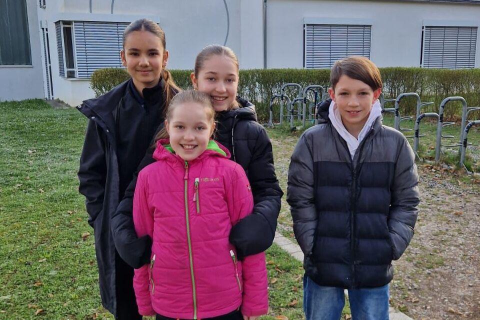 Vier Kinder stehen lächelnd vor dem Gebäude der Sportvereinigung FitKom, umgeben von Rasen und Radabstellplätzen.