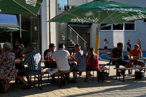Gruppe von Menschen sitzt an Tischen unter großen Markisen in einem Außenbereich, bei sonnigem Wetter.