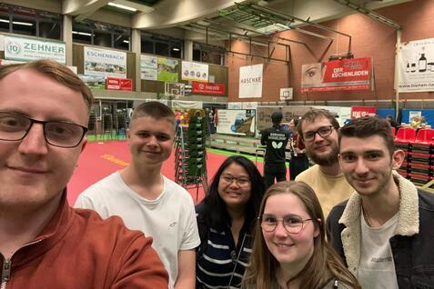 Gruppen-Selfie von fünf Personen in einer Sporthalle mit Tischtennis-Elementen im Hintergrund.
