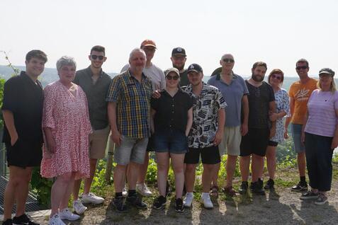 Gruppe von 16 Personen posiert auf einem Aussichtspunkt mit Weinbergen im Hintergrund und einem Verkaufsstand.