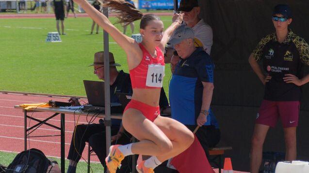 Leichtathletin in rotem Wettkampfanzug springt in den Sandkasten beim Weitsprung, Betreuer beobachten den Versuch.