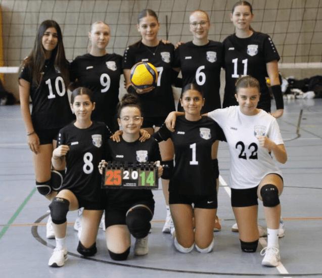 Gruppensfoto des Volleyballteams mit 10 Spielerinnen in schwarzen und weißen Trikots, einem Ball und einem Punktestand.