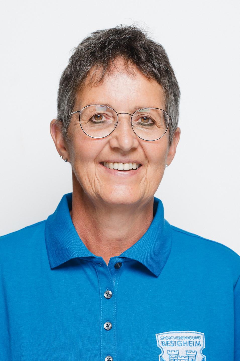 Portrait einer lächelnden Frau mit Kurzhaarfrisur, Brille und blauer Sportverein-Tunika vor weißem Hintergrund.