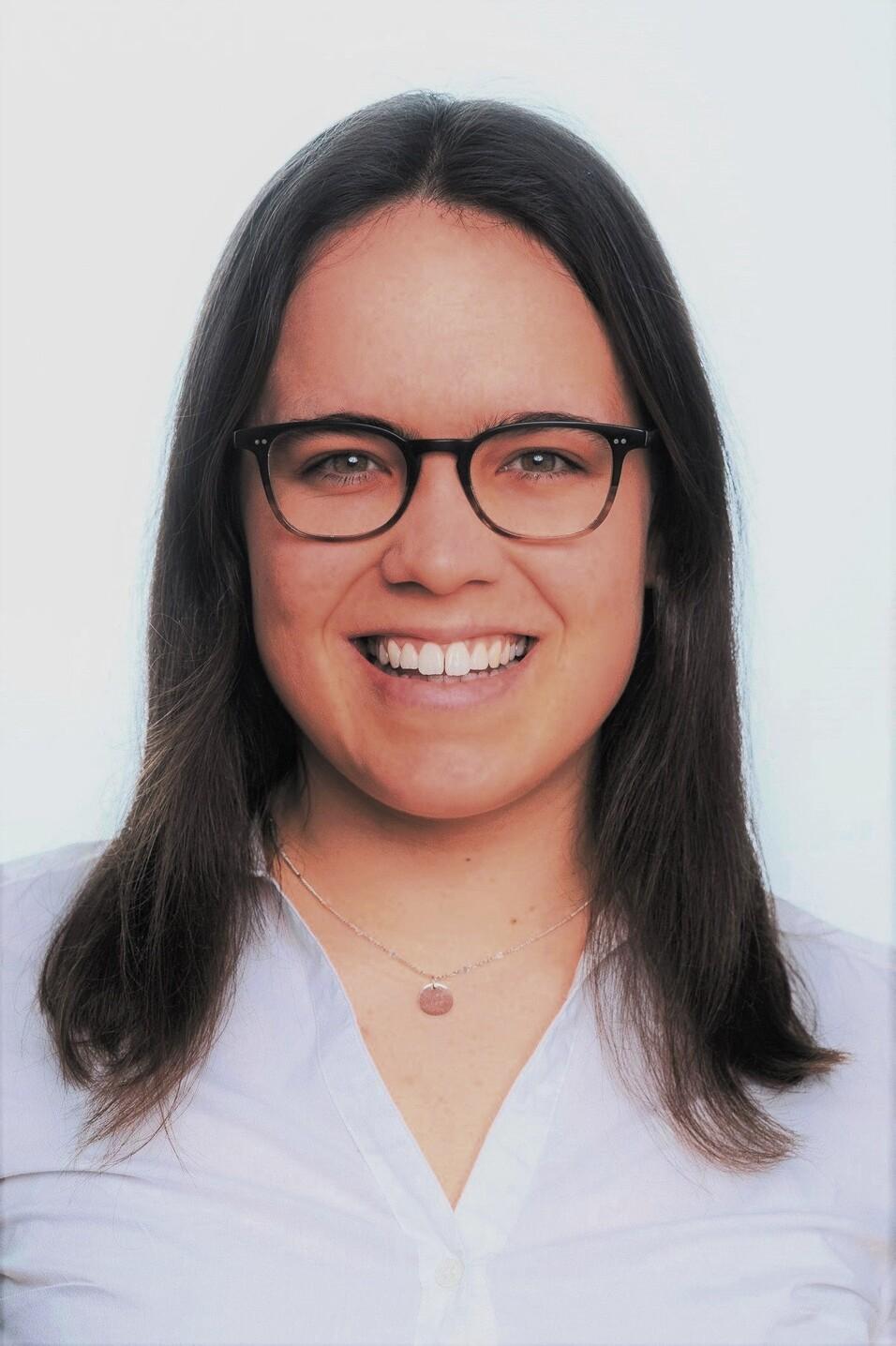 Lächelnde Frau mit langen Haaren und Brille, trägt ein hellblaues Hemd und eine filigrane Halskette, vor hellem Hintergrund.