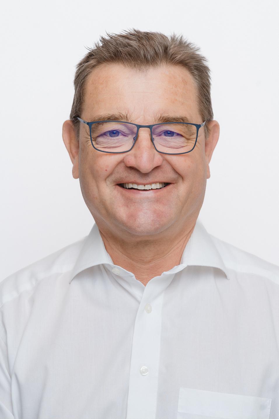 Lächelnder Mann mit Brille in weißem Hemd vor neutralem Hintergrund.