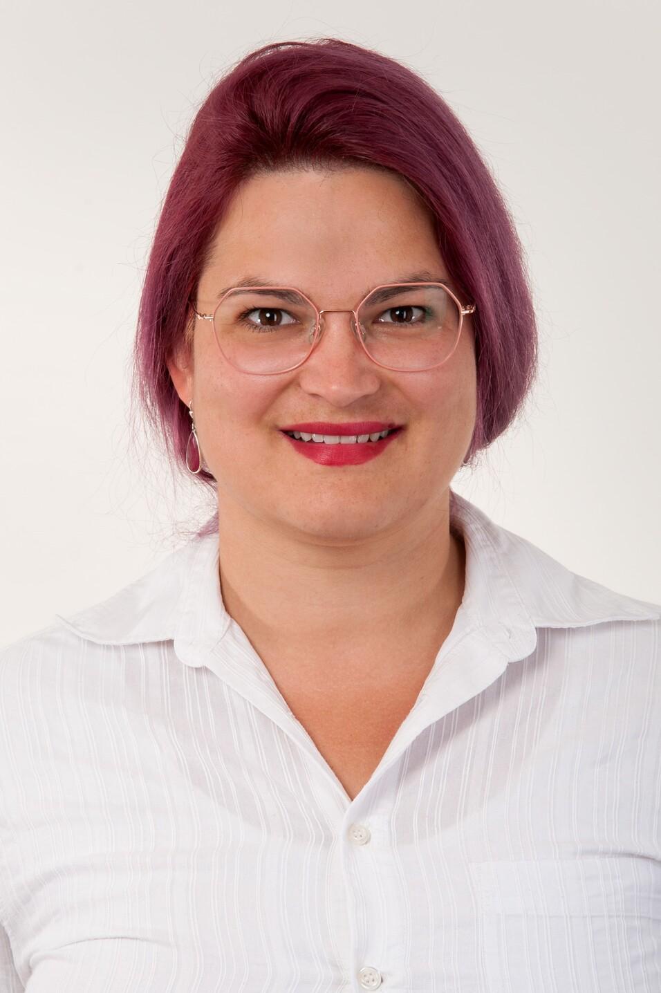 Porträt einer Person mit lila Haaren und Brille, die ein weißes, gestreiftes Hemd trägt, vor neutralem Hintergrund.