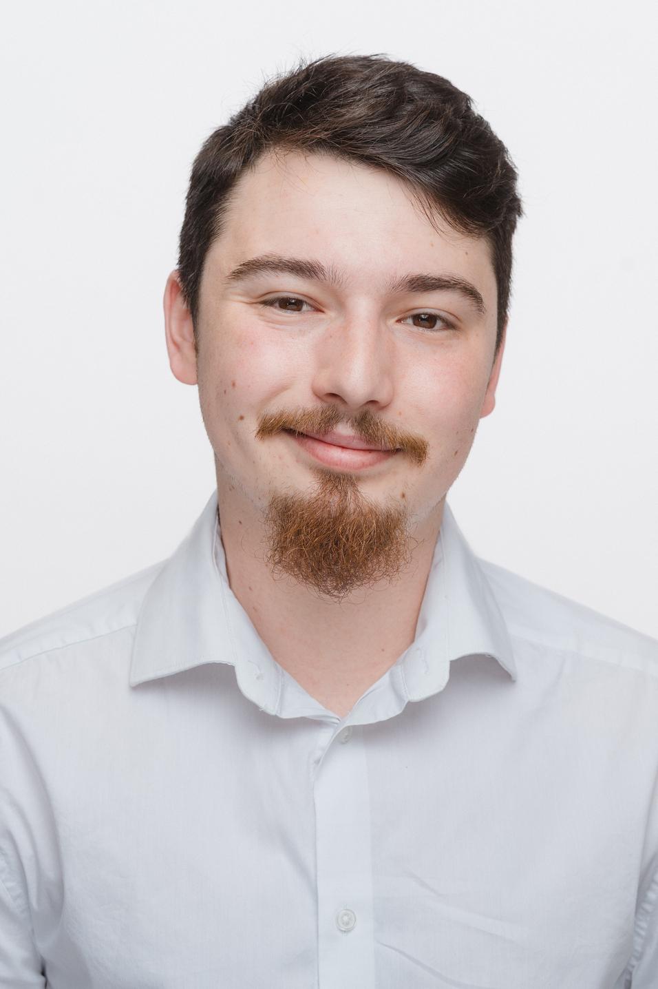 Porträt eines jungen Mannes mit Bart, der in einem weißen Hemd vor einem weißen Hintergrund lächelt.