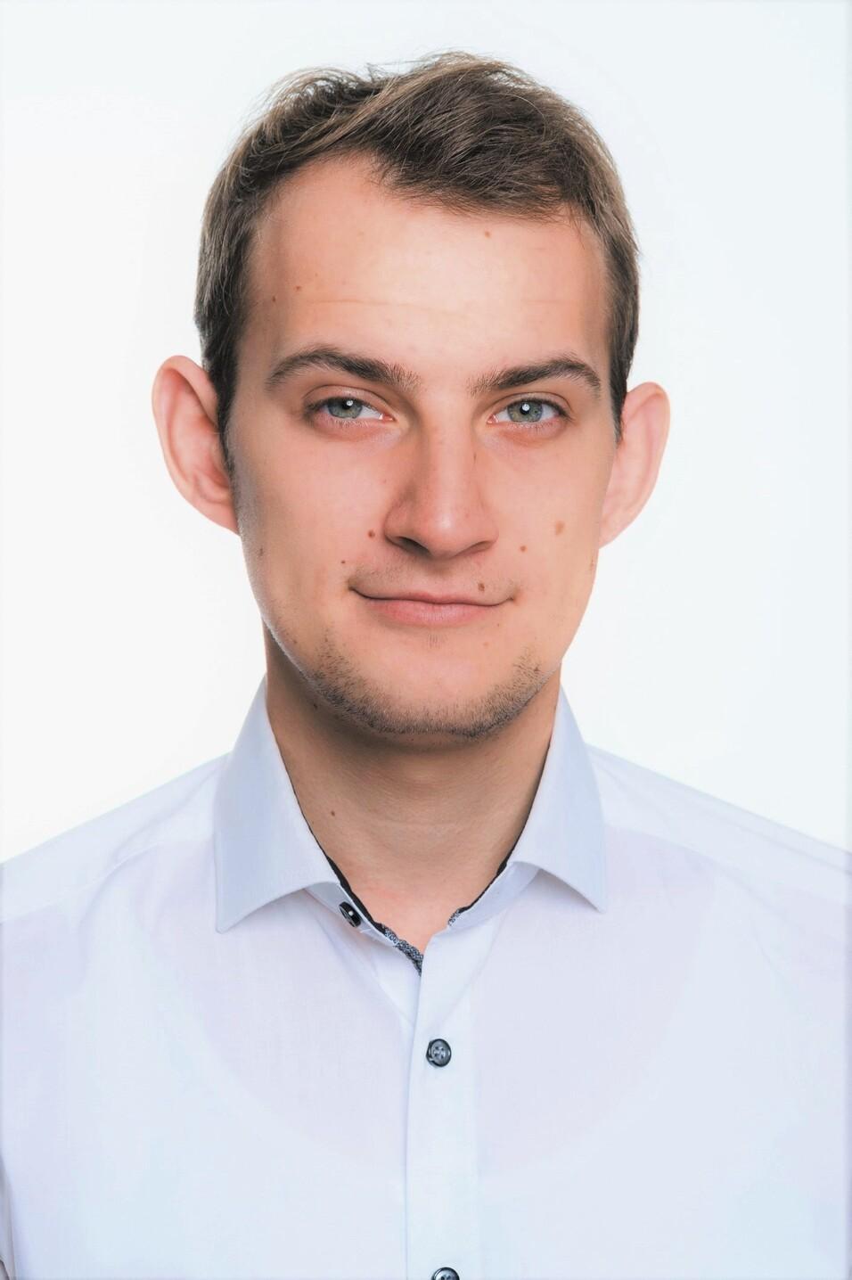 Porträt eines Mannes mit kurzen Haaren, blauen Augen und weißem Hemd vor einem hellen Hintergrund.