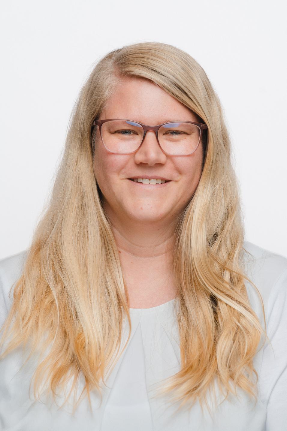 Porträt einer lächelnden Frau mit langen, blonden Haaren und Brille vor einem weißen Hintergrund.