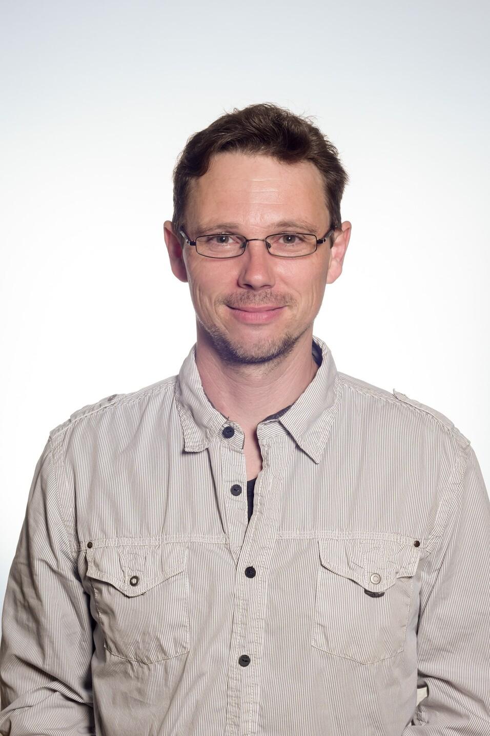 Porträt eines Mannes mit Brille in einem gestreiften Hemd vor einem hellen, einheitlichen Hintergrund.