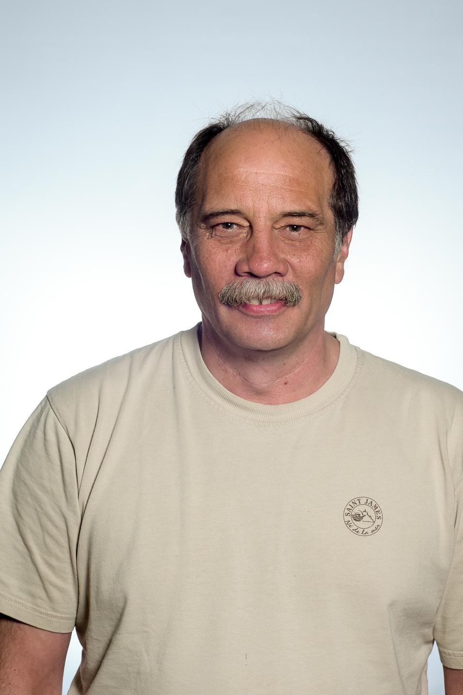 Porträt eines Mannes mit Schnurrbart, der ein beiges T-Shirt trägt, vor einem hellen Hintergrund.