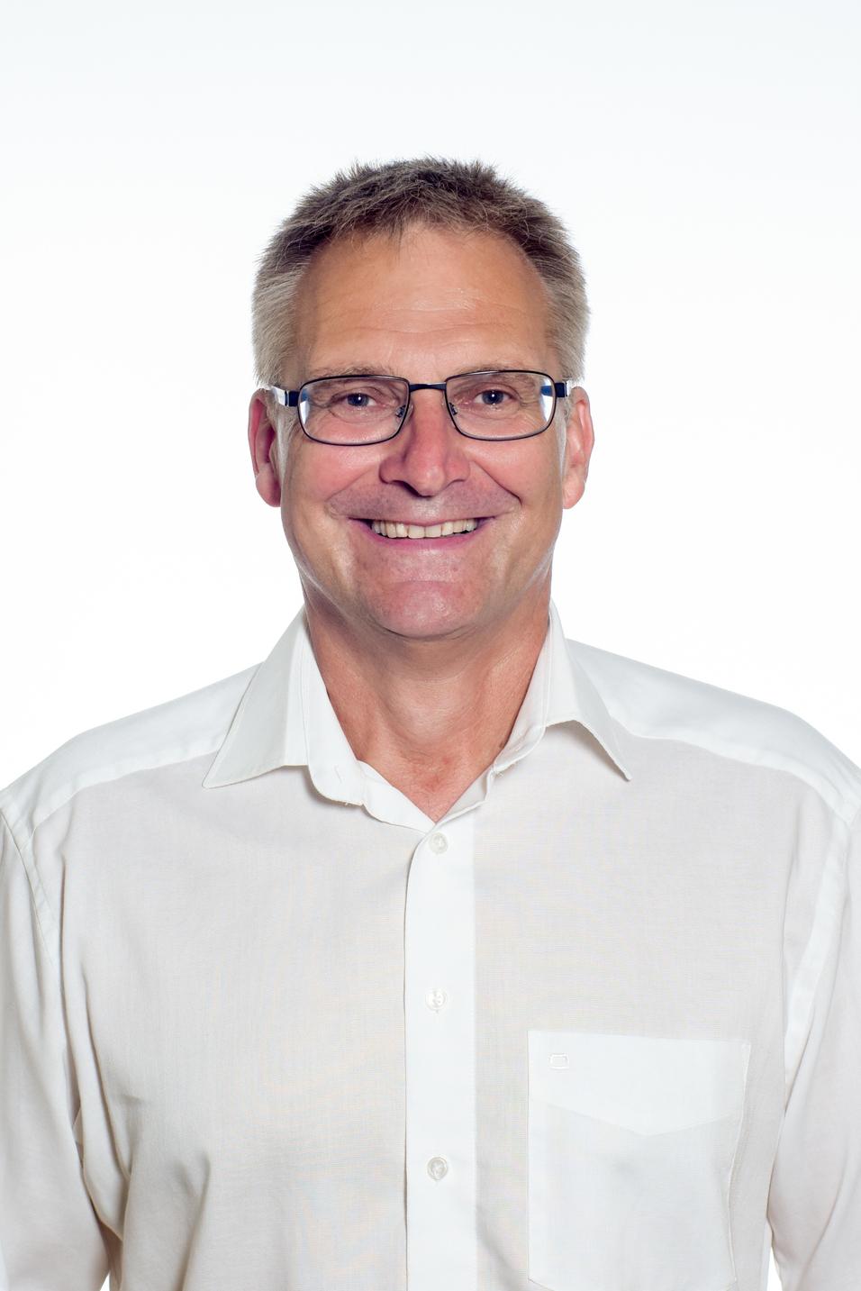 Lachenender Mann mit Brille in einem weißen Hemd, vor einem hellen Hintergrund. Kopf- und Oberkörperaufnahme.