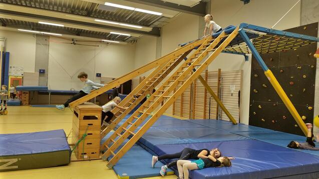 Kinder spielen in einer Turnhalle auf einem Holzgerüst mit Kletterelementen und Matten.