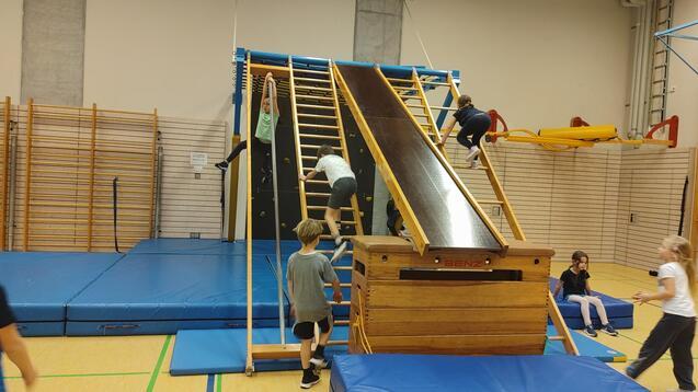 Kinder spielen in einer Turnhalle an einer Kletterwand mit Rutsche und Matten, während andere zuschauen.