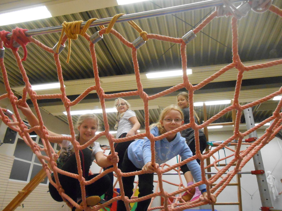 Vier Kinder spielen auf einem Klettergerüst mit einem Netz aus dicken, pinken Seilen in einer Turnhalle.