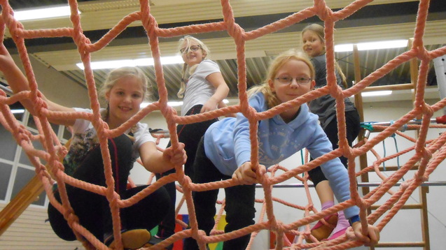 Vier Kinder spielen auf einem Klettergerüst mit einem Netz aus dicken, pinken Seilen in einer Turnhalle.