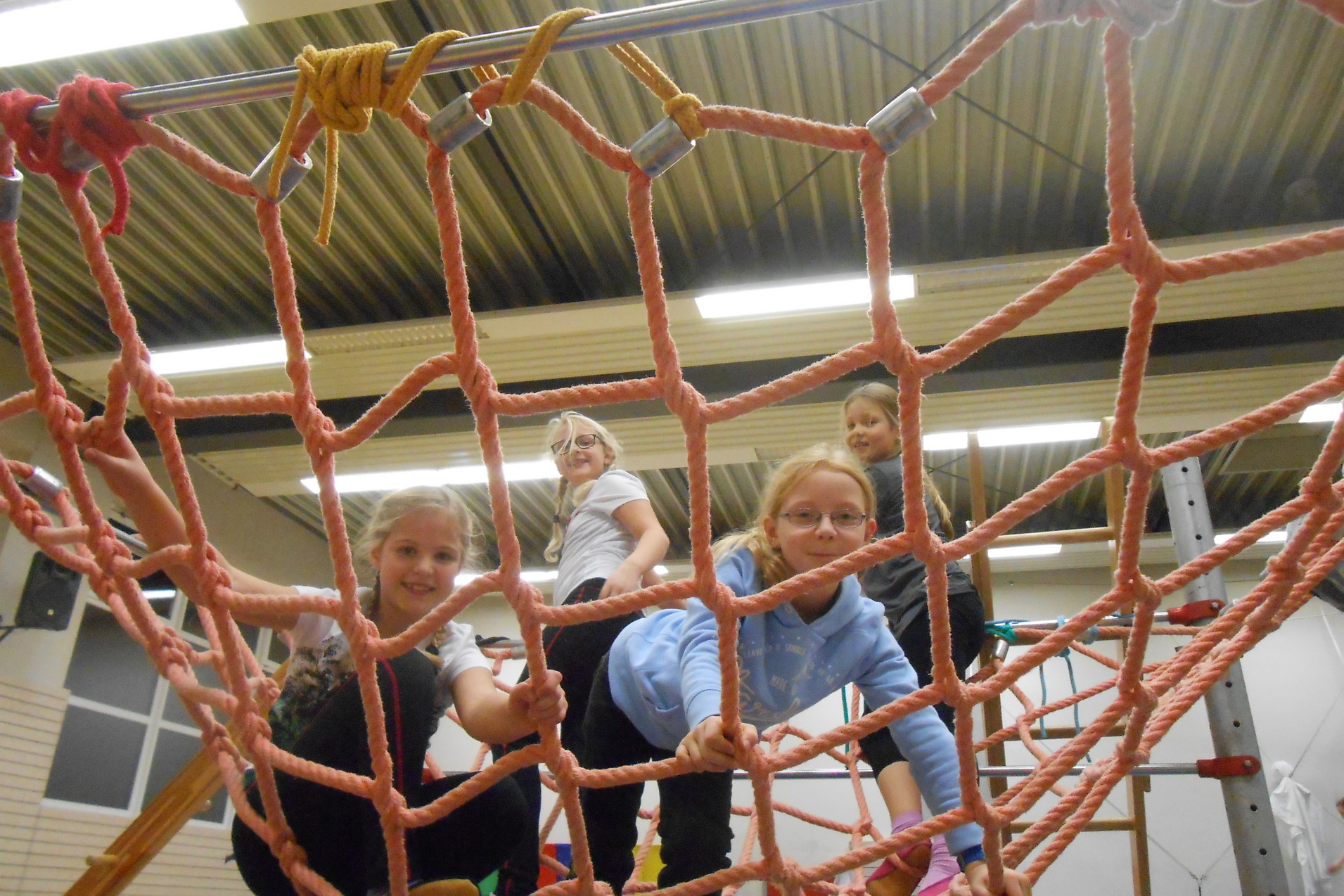 Vier Kinder spielen auf einem Klettergerüst mit einem Netz aus dicken, pinken Seilen in einer Turnhalle.