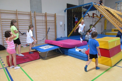 Kinder üben in einer Turnhalle mit Trampolin, Schaumstoffmatten und Klettergerüst. Aktivität und Bewegungsfreude sind sichtbar.