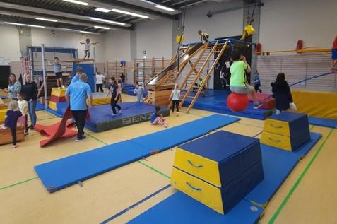 Kinder turnen in einer Sporthalle an verschiedenen Geräten wie Matten, einem Klettergerüst und einer Rutsche.