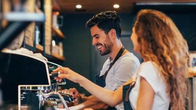 Zwei Personen arbeiten an einer Kaffeemaschine in einem Café, umgeben von weiteren Kaffeezubehör. Text über Stellenangebot.
