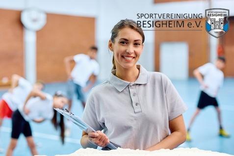 Lächelnde Frau mit Clipboard steht im Vordergrund, während im Hintergrund eine Gruppe in einer Sporthalle Übungen macht.