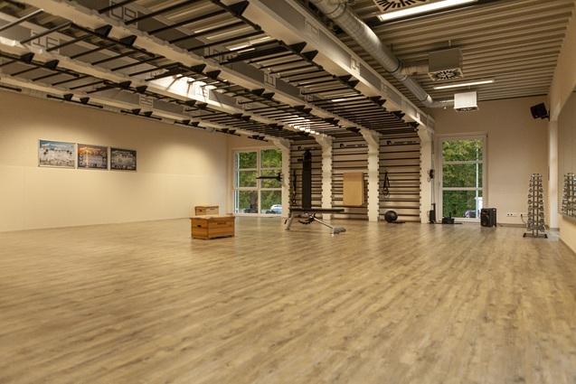 Großer, heller Fitnessraum mit Holzfußboden, Trainingsgeräten und Fenstern, die Blick ins Freie bieten.