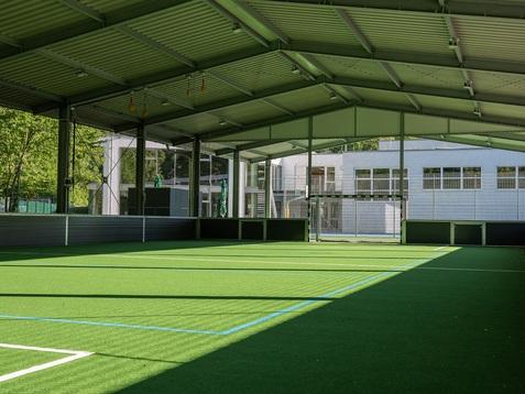 Heller, überdachter Sportplatz mit grünem Kunstrasen und Blick auf ein benachbartes Gebäude.
