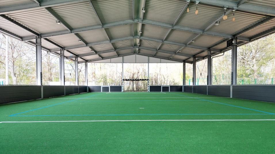 &Uuml;berdachte Sporthalle mit gr&uuml;nem Kunstrasen und Tor auf einer Seite, umgeben von Netzen und Fenstern.
