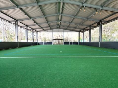 Überdachte Sporthalle mit grünem Kunstrasen und Tor auf einer Seite, umgeben von Netzen und Fenstern.