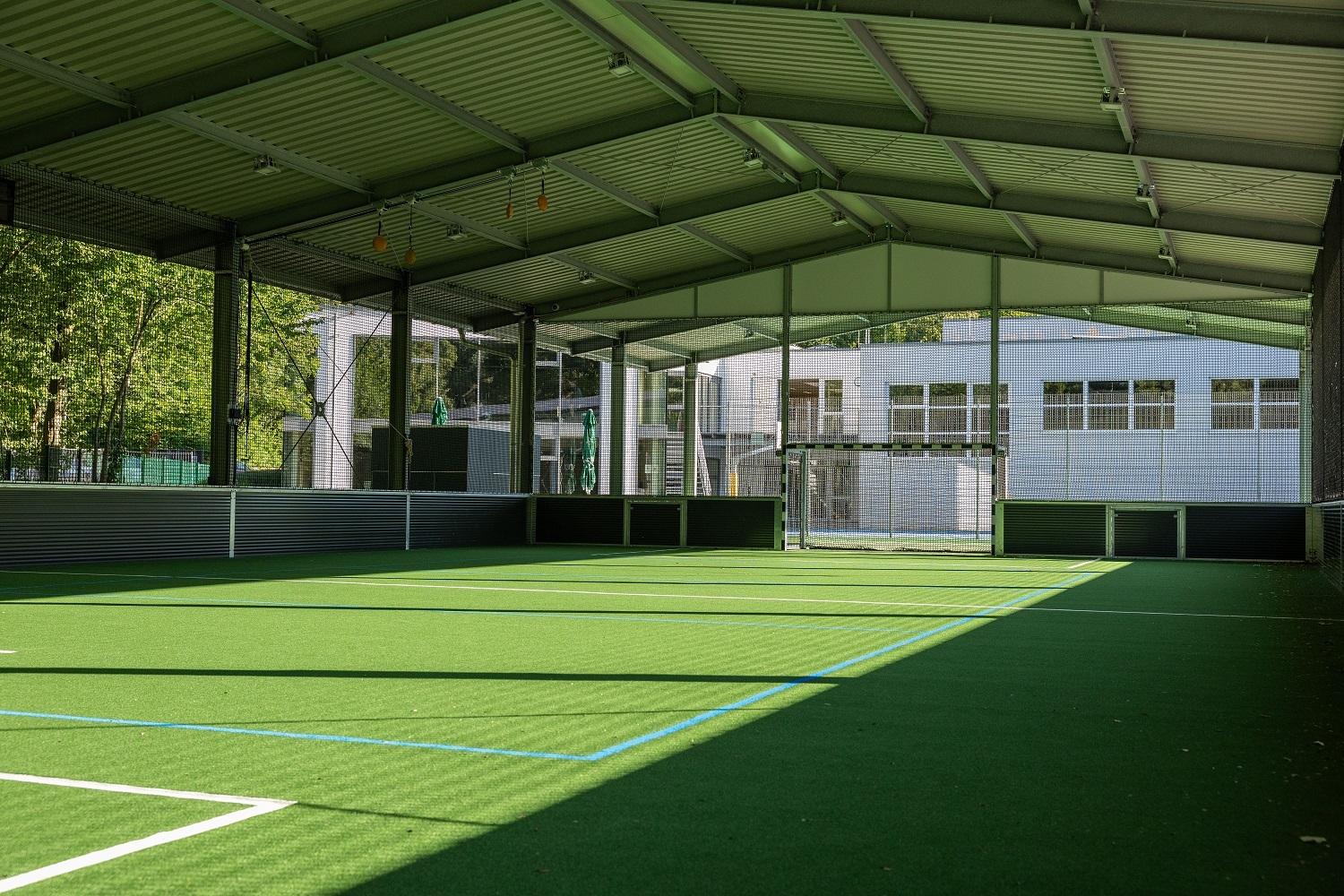 Heller, überdachter Sportplatz mit grünem Kunstrasen und Blick auf ein benachbartes Gebäude.