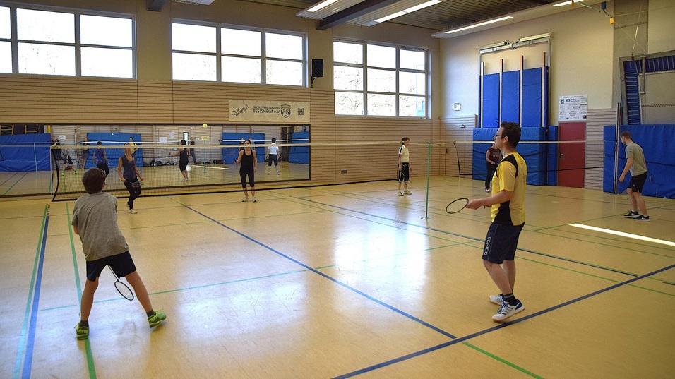 Sechs Personen spielen Badminton in einer Sporthalle mit Fensterfront und zwei Netzen.