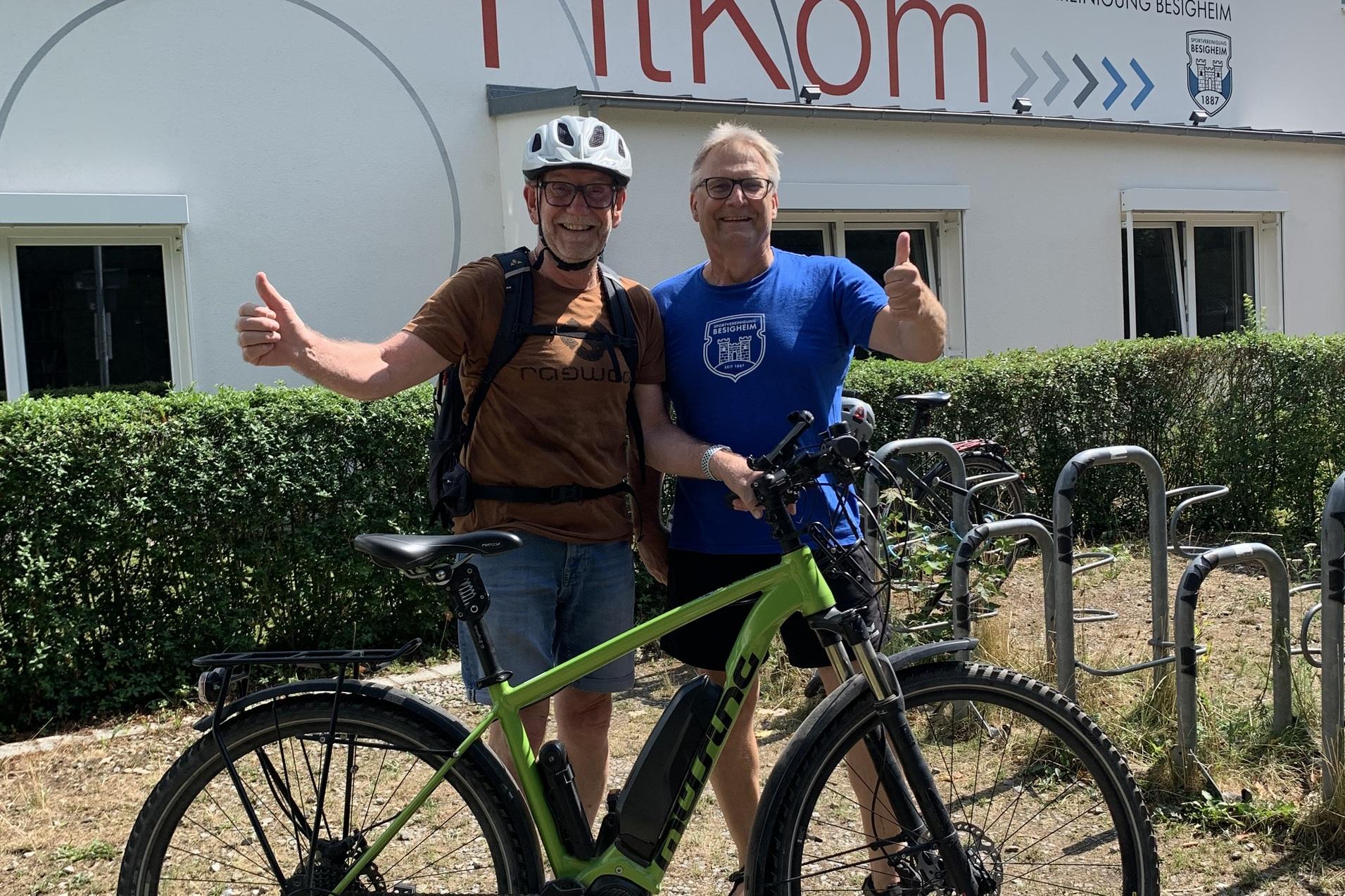 Zwei Männer stehen lächelnd vor einem Gebäude mit dem Schriftzug „FitKom“, neben einem grünen E-Bike.