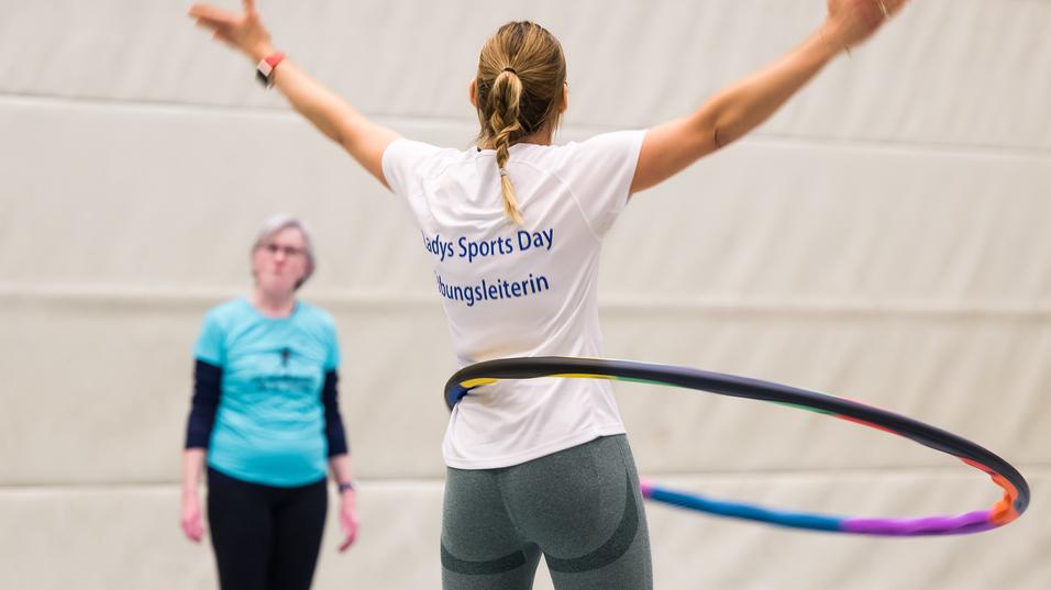 Sportveranstaltung: Frau mit Hula-Hoop-Reifen, im Hintergrund eine weitere Teilnehmerin, in einer Sporthalle.