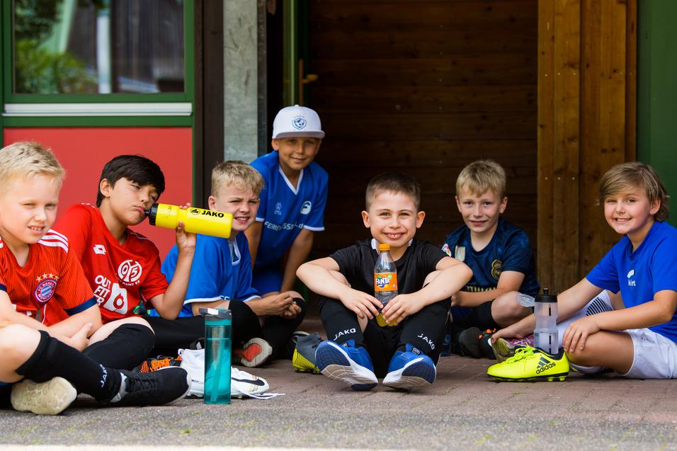 Sechs Kinder sitzen in Sportkleidung auf dem Boden, lächeln und halten Trinkflaschen und Snacks.