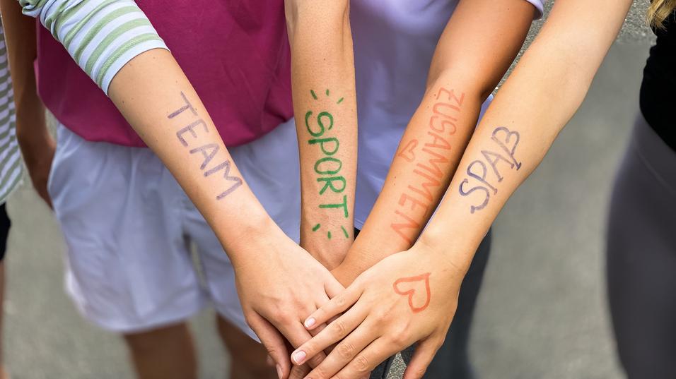 Vier Arme liegen übereinander, mit farbiger Schrift: „TEAM“, „SPORT“, „ZUSAMMEN“, „SPASS“ auf der Haut.
