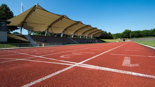 Nahaufnahme einer roten Laufbahn mit Startlinien und einem überdachten Zuschauerbereich im Hintergrund.