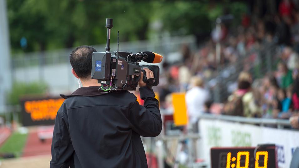 Kameramann filmt bei einer Sportveranstaltung, Hintergrund zeigt eine Zuschauertribüne und eine Zeitmessanzeige.