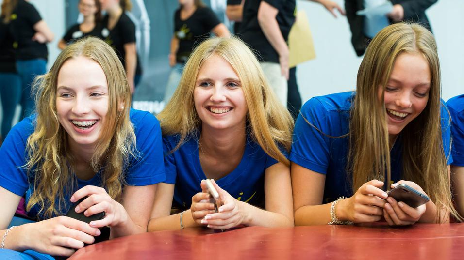 Drei lachende Jugendliche in blauen T-Shirts liegen auf einem Tisch und schauen auf ihre Handys. Hintergrund mit weiteren Personen.
