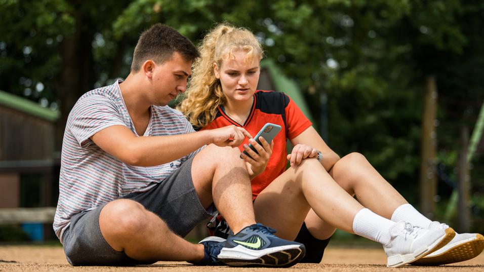 Zwei Jugendliche sitzen auf dem Boden und betrachten gemeinsam ein Smartphone. Ein Mädchen trägt ein rotes sportliches Oberteil.