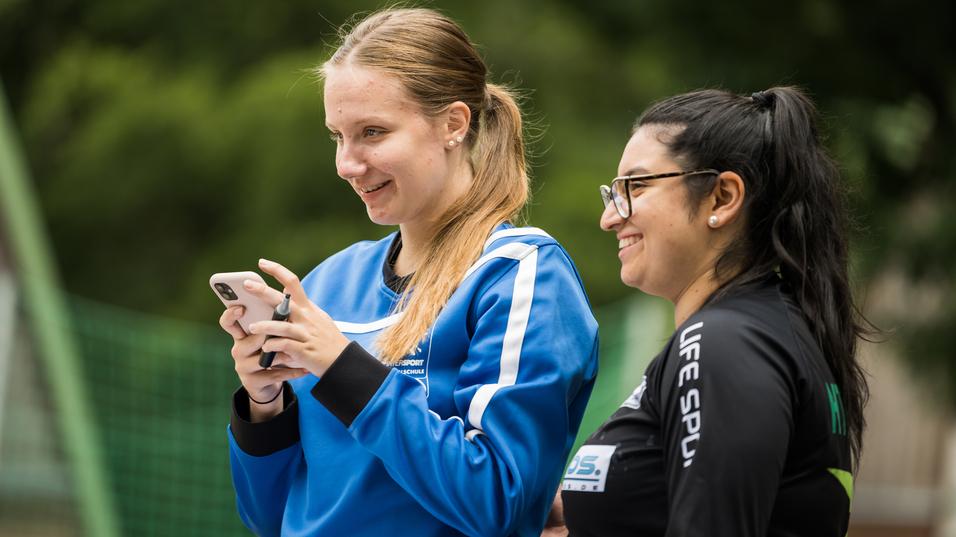 Zwei Frauen lächeln, während eine auf ihrem Smartphone tippt, umgeben von grüner Natur und Sportgeräten im Hintergrund.