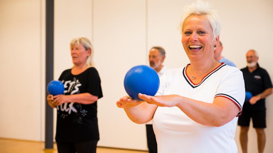 Gruppe älterer Menschen bei einer Übung mit blauen Bällen in einem hellen Raum. Fokus auf lächelnde Frau vorne.