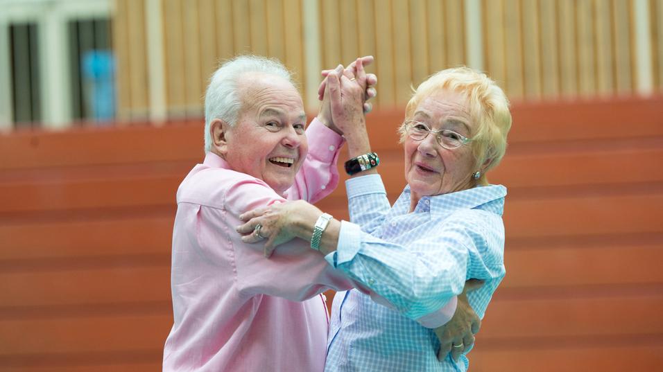 Ein älteres Paar tanzt lächelnd in einer Turnhalle, Kleidung in sanften Farben. Holzpaneele im Hintergrund.