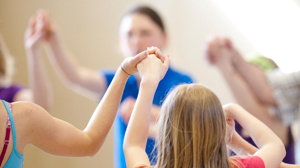 Gruppe von Kindern hält sich in einem gemeinsamen Kreis an den Händen, während sie in einer Sporthalle aktiv sind.