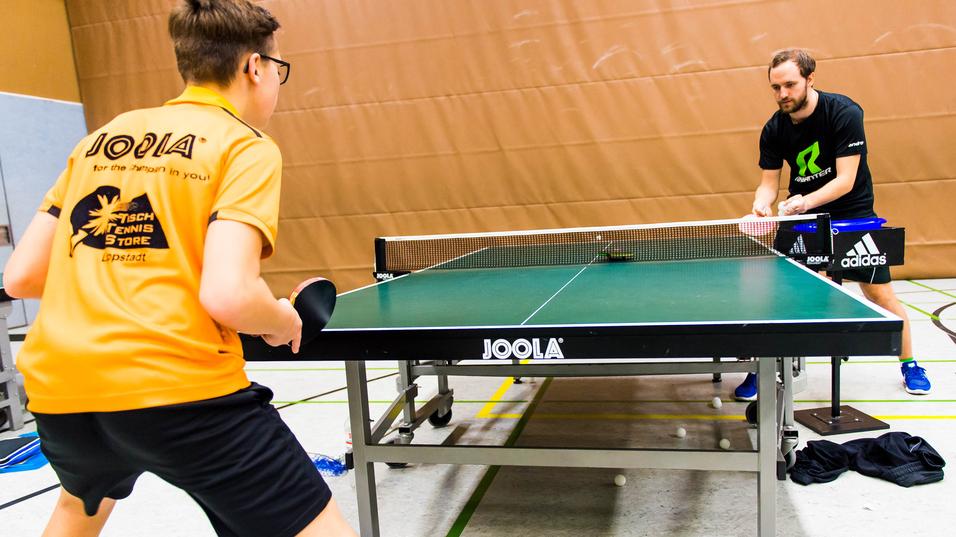Zwei Tischtennisspieler trainieren an einem Tischtennistisch in einer Sporthalle, mehrere Bälle liegen auf dem Boden.