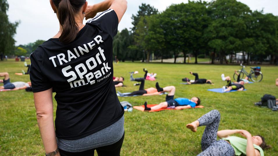 Trainerin mit schwarzem Shirt leitet Sportübungen im Park; Teilnehmer liegen auf Matten und trainieren im Hintergrund.