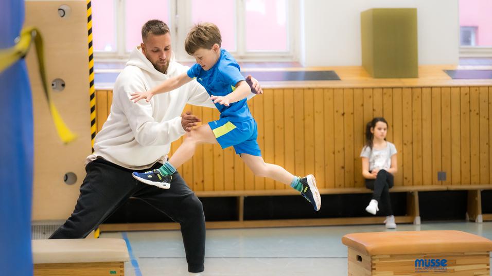 Ein junger Junge springt mit Hilfe eines Trainers über eine Trainingsbank in einer Turnhalle. Ein Mädchen beobachtet.